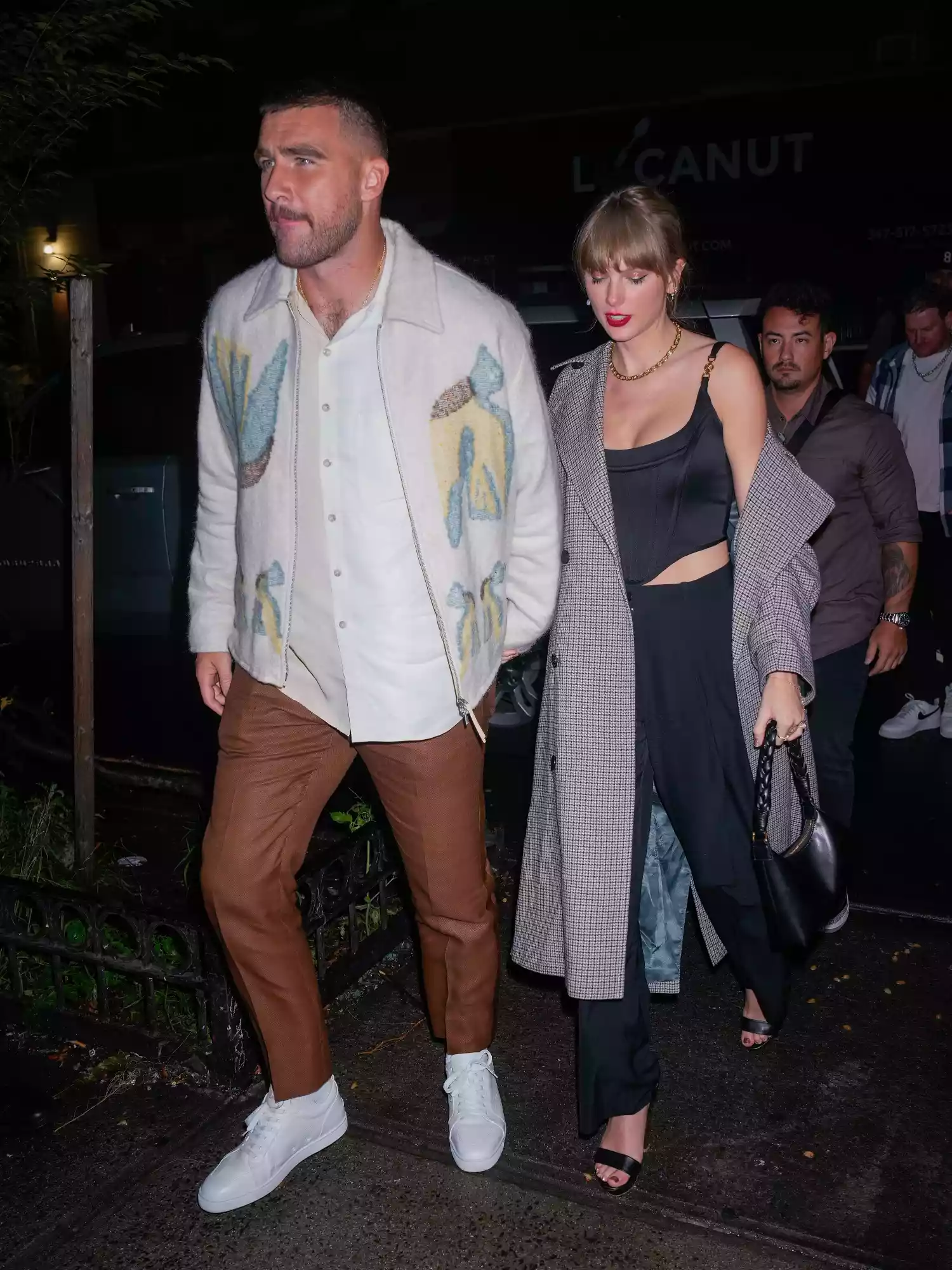 Taylor Swift wearing black strappy heels, black pants, black corset, gray trench coat, and Aupen handbag while holding hands with Travis Kelce