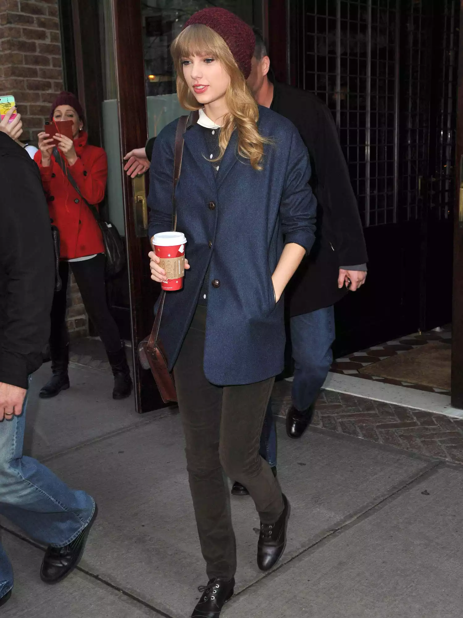 Taylor Swift wearing a maroon beanie, blue coat, green pants, black flats, and holding coffee cup