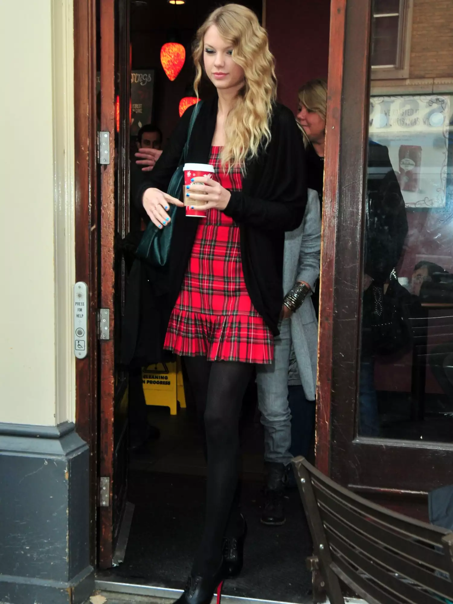 Taylor Swift wearing a red gingham dress, black coat, black shoes, and holding coffee cup